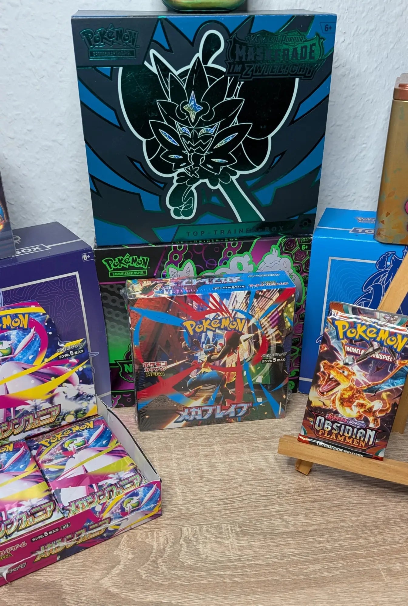 Eine Sammlung verschiedener Pokémon-Sammelkartenprodukte auf einem Tisch, darunter eine Top-Trainer-Box 'Maskerade im Zwielicht', eine japanische Booster-Box und Booster-Packs.