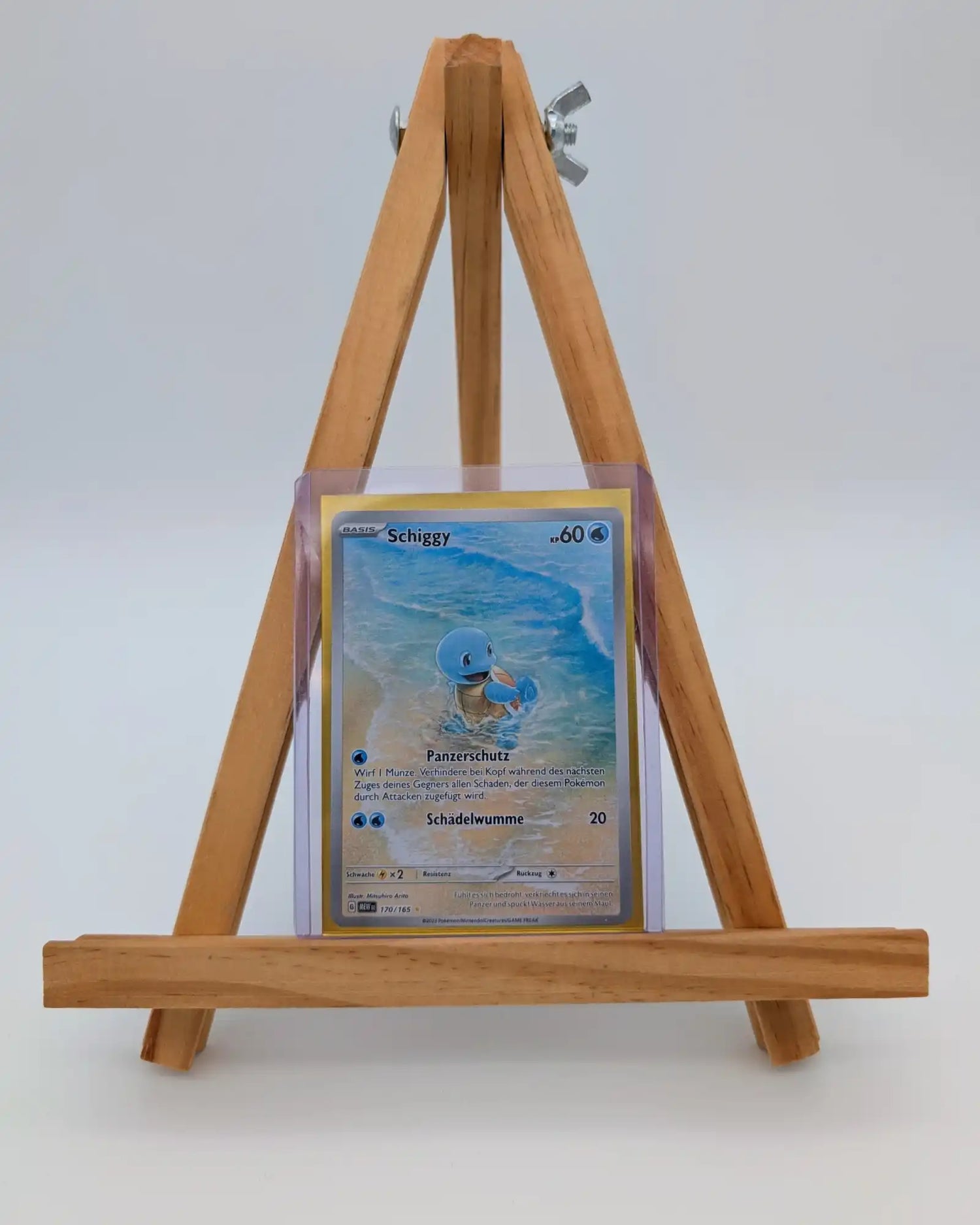 Eine Pokémon-Sammelkarte des Charakters Schiggy, die in einer Schutzhülle auf einer kleinen Holzstaffelei präsentiert wird. Das Bild ist vor einem schlichten, weißen Hintergrund aufgenommen.